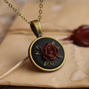 Può includere: Una collana con pendente in stile vintage con catena in bronzo. Il pendente presenta uno sfondo nero con un motivo a rosa rossa in rilievo e una cornice color bronzo. La collana è appoggiata su una lettera vintage.