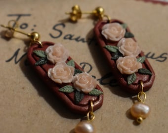 May include: A pair of handcrafted earrings featuring a rectangular, reddish-brown base adorned with three peach-colored rose accents and green leaves. Each earring has a dangling, pear-shaped pearl. The earrings are set against a vintage paper background.