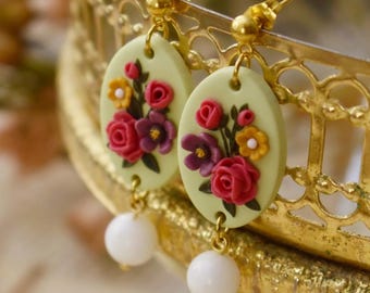 May include: A pair of floral earrings with a pale green oval base and a cluster of red, pink, and yellow flowers. The earrings have a white pearl dangle and gold hooks.