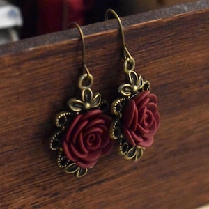 Può includere: Un paio di orecchini pendenti con un design a rosa bordeaux. Le rose sono realizzate in pasta polimerica e sono circondate da accenti metallici dorati.