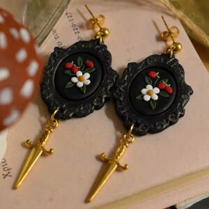May include: A pair of black framed earrings with a floral design and gold dagger pendants. Each earring features a black oval with a white flower and red berries. The earrings have gold-colored posts.