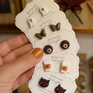 Può includere: Cinque paia di orecchini a lobo su cartoncini con il marchio melissaki, raffiguranti fantasmi bianchi, farfalle monarca arancioni e nere, ricci marroni, tazze di caffè bianche con zucche e teste di gatto nere decorate con rose rosse.