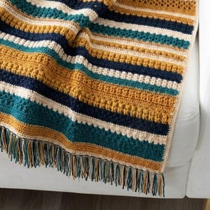 May include: A crocheted throw blanket featuring horizontal stripes in mustard yellow, navy blue, teal, and cream. The blanket displays various textured stitch patterns, including bobble stitches, and is finished with a fringe of multi-colored yarn.