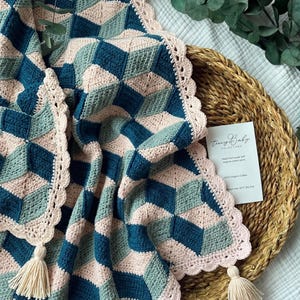 May include: A crochet blanket featuring a geometric pattern of dark teal, sage green, and pale pink diamond shapes. The blanket has a scalloped pale pink border with cream-colored tassels at the corners and includes a white product information card.