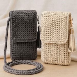 May include: Two rectangular crochet crossbody bags, one in dark charcoal gray and one in cream, each featuring a flap closure, a long matching cord strap, and a decorative tassel attached to a gold-toned metal clasp on the side of the bag.