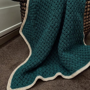 May include: A deep teal crochet blanket featuring a textured basketweave pattern throughout the main body. The blanket is finished with a solid cream-colored border that frames the edges, providing a distinct contrast to the dark teal yarn.
