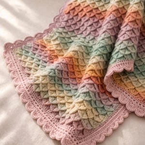 May include: A crochet blanket featuring a textured crocodile stitch pattern in a gradient of pastel colors including pink, yellow, green, and orange. The blanket is finished with a scalloped border in a solid light pink yarn throughout the edges.