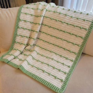 May include: A crocheted blanket featuring a white base with horizontal rows of light green bobble stitches. The blanket is finished with a wide, textured light green border that frames the entire piece, creating a consistent pattern throughout.