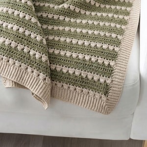 May include: A crocheted blanket featuring alternating horizontal stripes of sage green and cream yarn. The design incorporates rows of textured bobble stitches between solid crochet sections, finished with a ribbed cream border along the edges.