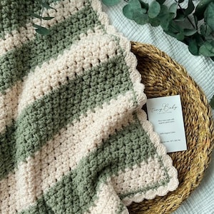 May include: A crocheted blanket featuring alternating horizontal stripes of sage green and cream yarn. The blanket has a scalloped edge border and is accompanied by a small white rectangular card with the text Tiny Baby Collections.