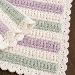 Könnte beinhalten: Eine handgehäkelte Decke mit abwechselnden horizontalen Streifen in Creme, zartem Lavendel und Salbeigrün. Das Design umfasst strukturierte Noppenstichreihen und einen dekorativen, bogenförmigen Randabschluss aus cremefarbenem Garn.