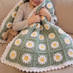 May include: A handmade crochet blanket featuring a repeating pattern of sage green square motifs, each centred with a white and yellow daisy flower. The blanket is finished with a white scalloped border and is accompanied by a small brown plush toy.