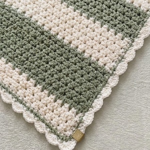 May include: A crocheted blanket featuring alternating wide stripes of sage green and cream yarn. The textile displays a consistent textured stitch pattern throughout, finished with a delicate scalloped edge border and a small rectangular tan tag.