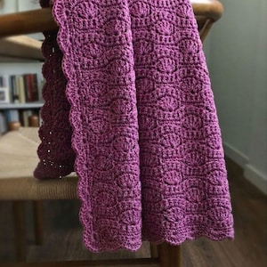May include: A deep magenta crochet blanket featuring a repeating shell stitch pattern throughout the fabric. The edges of the textile are finished with a scalloped border, creating a decorative and textured appearance across the entire surface.