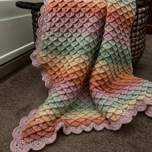 May include: A crochet blanket featuring a textured scale pattern in a gradient of pastel colors including pink, peach, yellow, and light green. The blanket is finished with a scalloped light pink border along the edges, creating a soft appearance.