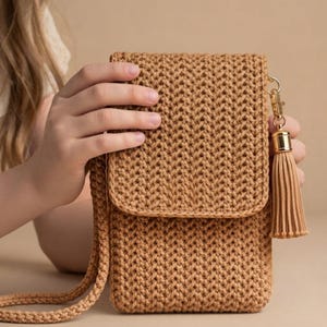 Puede incluir: Un bolso bandolera para teléfono hecho de ganchillo color canela con un patrón de punto de espiga texturizado, una solapa plegable y una correa larga a juego. Un cierre de metal dorado sujeta una borla decorativa de hilo color canela.