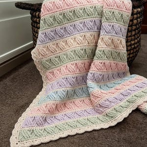 May include: A hand-knitted blanket featuring alternating horizontal stripes in pastel pink, mint green, lavender, and light blue. The blanket displays a textured cable knit pattern throughout and is finished with a scalloped cream-colored border.