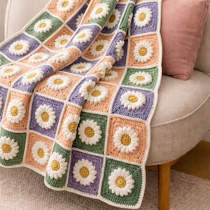 Könnte beinhalten: Eine Häkeldecke aus quadratischen Patches mit einem dreidimensionalen weißen Gänseblümchenmotiv und gelber Mitte. Die Quadrate sind in einem Rastermuster mit abwechselnden Hintergrundfarben in Salbeigrün, hellem Pfirsich und Lavendel angeordnet.