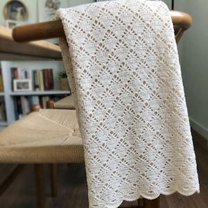 May include: An off-white crochet blanket featuring a repeating diamond pattern with openwork details and a scalloped edge. The textile is draped over a wooden chair, showcasing the intricate needlework and the soft, light-colored yarn texture.