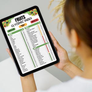 May include: A person holds a tablet displaying a "Fruits Glycemic Index" chart. The chart lists various fruits and juices with their glycemic index values, color-coded for low, medium, and high GI levels. The tablet has a black frame and is held against a white background.