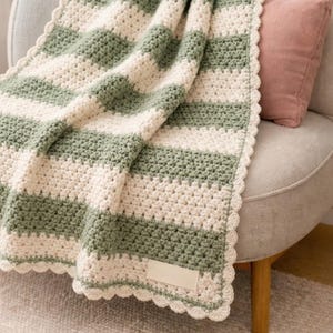 Puede incluir: Una manta de ganchillo con rayas horizontales alternas de hilo verde salvia y crema. La manta tiene un borde festoneado y una etiqueta rectangular de tela blanca en blanco cosida en la esquina inferior de la tela texturizada.