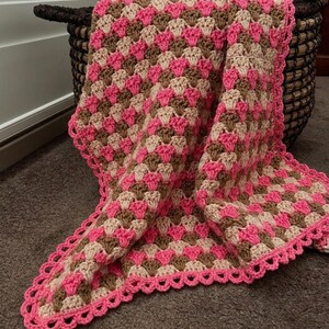 May include: A crocheted blanket featuring a repeating shell stitch pattern in alternating rows of bright pink, light beige, and medium brown yarn. The edges of the blanket are finished with a decorative scalloped border in bright pink yarn.