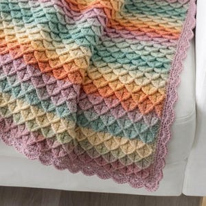 Könnte beinhalten: Eine Häkeldecke mit einem strukturierten Krokodilstichmuster in einem Verlauf aus Pastellfarben wie Rosa, Gelb, Orange und Hellgrün. Die Decke ist mit einer gewellten, malvenfarbenen Bordüre an der sichtbaren Kante versehen.