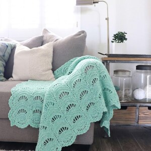 May include: A mint green crochet blanket featuring a repeating fan or shell stitch pattern with a scalloped edge. The textile is draped over a gray sofa, showcasing the intricate yarn work and the soft, light-colored texture of the handmade fabric.
