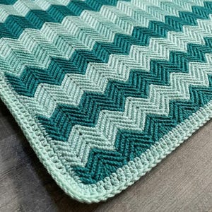 Op de afbeelding: Een gehaakte deken met een herhalend chevronpatroon in afwisselende tinten donkerblauwgroen, middenblauwgroen en licht muntgroen. Het textiel heeft een getextureerd, geribbeld uiterlijk met een effen licht muntgroene rand langs de zijkant.