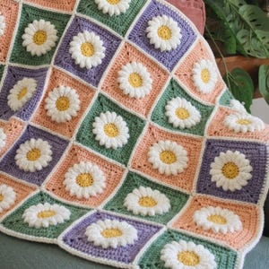 May include: A crochet blanket made of square patches in shades of sage green, lavender, and peach. Each square features a raised, three-dimensional white daisy with a yellow centre. The squares are joined together to form a repeating floral pattern.