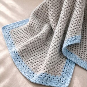 Peut inclure: Une couverture en crochet texturée et douce, dotée d'un centre gris clair avec un motif de points en relief. La couverture est finie par une bordure décorative en crochet bleu clair qui encadre tout le contour du tissu gris.