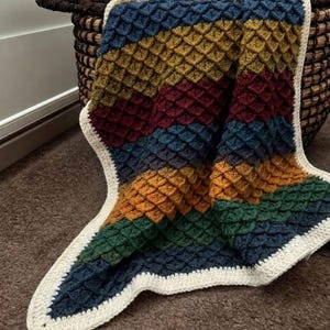 Può includere: Una coperta all'uncinetto caratterizzata da un motivo a punto waffle testurizzato a strisce orizzontali blu, giallo senape, bordeaux e verde scuro. La coperta è rifinita con un bordo color crema a tinta unita che incornicia il pezzo.