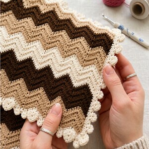 May include: A rectangular crochet piece featuring a chevron pattern in alternating stripes of dark brown, tan, and cream. The edges are finished with a decorative cream-coloured scalloped border consisting of small, rounded crochet clusters.