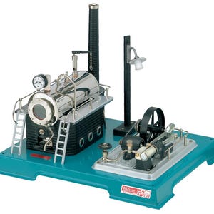 May include: A silver and black model steam engine with a blue base. The engine has a boiler, a piston, and a flywheel. It also has a small lamp on a post. The engine is on a blue base with the word "Wilesco" printed on it.