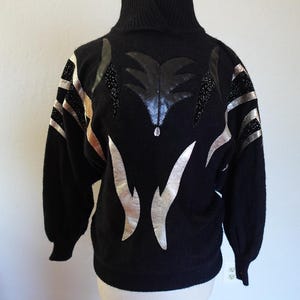 Könnte beinhalten: Schwarzer Rollkragenpullover mit silbernen und goldenen Akzenten. Der Pullover hat ein einzigartiges Design mit silbernen flammenartigen Formen und goldenen Streifen auf den Ärmeln. Ein schwarzes Paillettenmuster befindet sich in der Mitte der Brust. Der Pullover wird auf einer Holzpuppe präsentiert.
