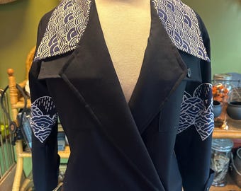 May include: Black jacket with a unique design. The jacket features a contrasting collar and sleeve details with a white and navy blue wave pattern. The jacket has a double-breasted closure with two black buttons.
