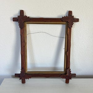May include: A rectangular wooden picture frame featuring a dark brown finish with carved leaf-shaped corner accents and a thin gold-toned inner border. A thin metal hanging wire is attached across the upper interior section of the frame.