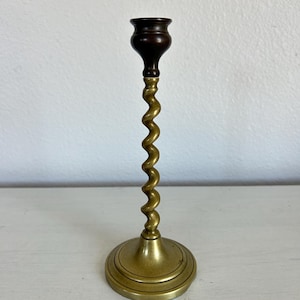 May include: A candlestick holder featuring a circular brass base, a central spiraled brass stem, and a dark brown wooden candle cup at the top. The metal components have a polished finish, while the wooden top provides a contrasting dark tone.