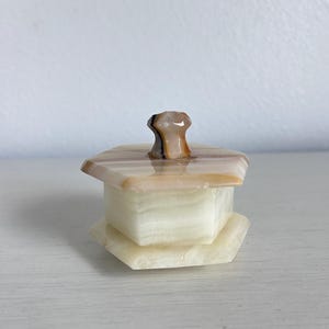May include: A small, hexagonal onyx box with a lid. The box is primarily off-white with bands of tan and brown. The lid has a small, rounded knob. The box sits on a white surface.