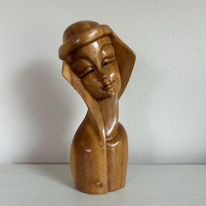 May include: A wooden sculpture of a woman's head and shoulders. The sculpture is carved from light brown wood and features a hat and a draped collar. The woman's face has closed eyes and a slight smile. The sculpture is displayed on a white surface.