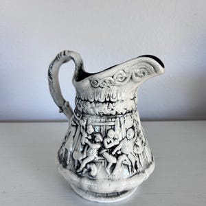 May include: A small, white and black ceramic pitcher with a detailed relief design. The pitcher features a handle and a spout, with a decorative band around the top and a scene of figures engaged in activities around the body.