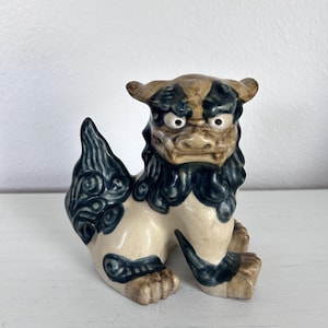 May include: A ceramic Shisa dog statue, a traditional Ryukyuan guardian figure. The statue features a cream-colored body with dark blue and brown accents. The Shisa is depicted in a seated position with a fierce expression.