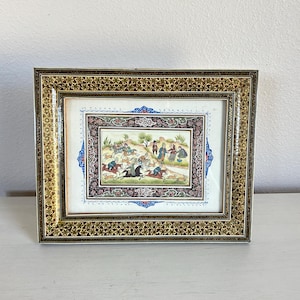 May include: A rectangular framed painting featuring a detailed miniature scene of figures on horseback and on foot in a landscape. The frame has a dark brown and gold intricate geometric pattern, surrounding a central image with a floral border.