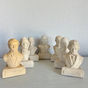 May include: A collection of seven off-white and light tan resin busts of historical composers. Each bust sits on a triangular base with the name of the composer and their birth and death years embossed on the front, including Wagner and Beethoven.