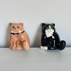May include: Two ceramic cat figurines. One is a light orange cat with a black and white polka dot bow tie and green eyes. The other is a black and white tuxedo cat with a white chest, white paws, and green eyes. Both cats are in a seated position.