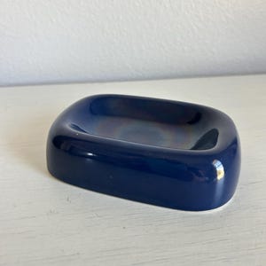 May include: A rectangular soap dish with rounded corners and a smooth, glossy finish. The object features a deep navy blue color with a subtle, iridescent metallic sheen visible in the center basin, creating a reflective and polished appearance.