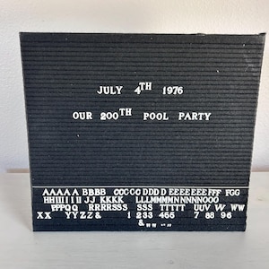 May include: A black felt letter board with white lettering. The text reads "JULY 4TH 1976 OUR 200TH POOL PARTY." Additional letters and numbers are displayed below. The board is rectangular and sits on a white surface.