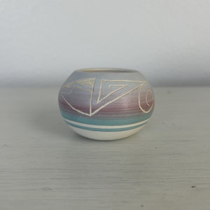 May include: A small, round ceramic vase with a light blue, purple, and teal color scheme. The vase features a geometric design in white. A white stripe encircles the base. The vase is sitting on a white surface.