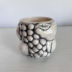 May include: A small, off-white ceramic cup with a grape cluster design. The cup's exterior features a textured pattern of grape-like spheres, with dark gray detailing. The interior is a lighter shade, and the cup has a smooth, rounded rim.