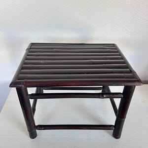 May include: A small, square bamboo stool with a dark reddish-brown finish. The top is made of parallel bamboo slats, and the legs are angled for stability. The stool is designed with a minimalist aesthetic.